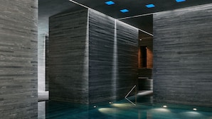 A modern indoor swimming pool with a minimalist design, featuring dark walls and blue lighting accents.