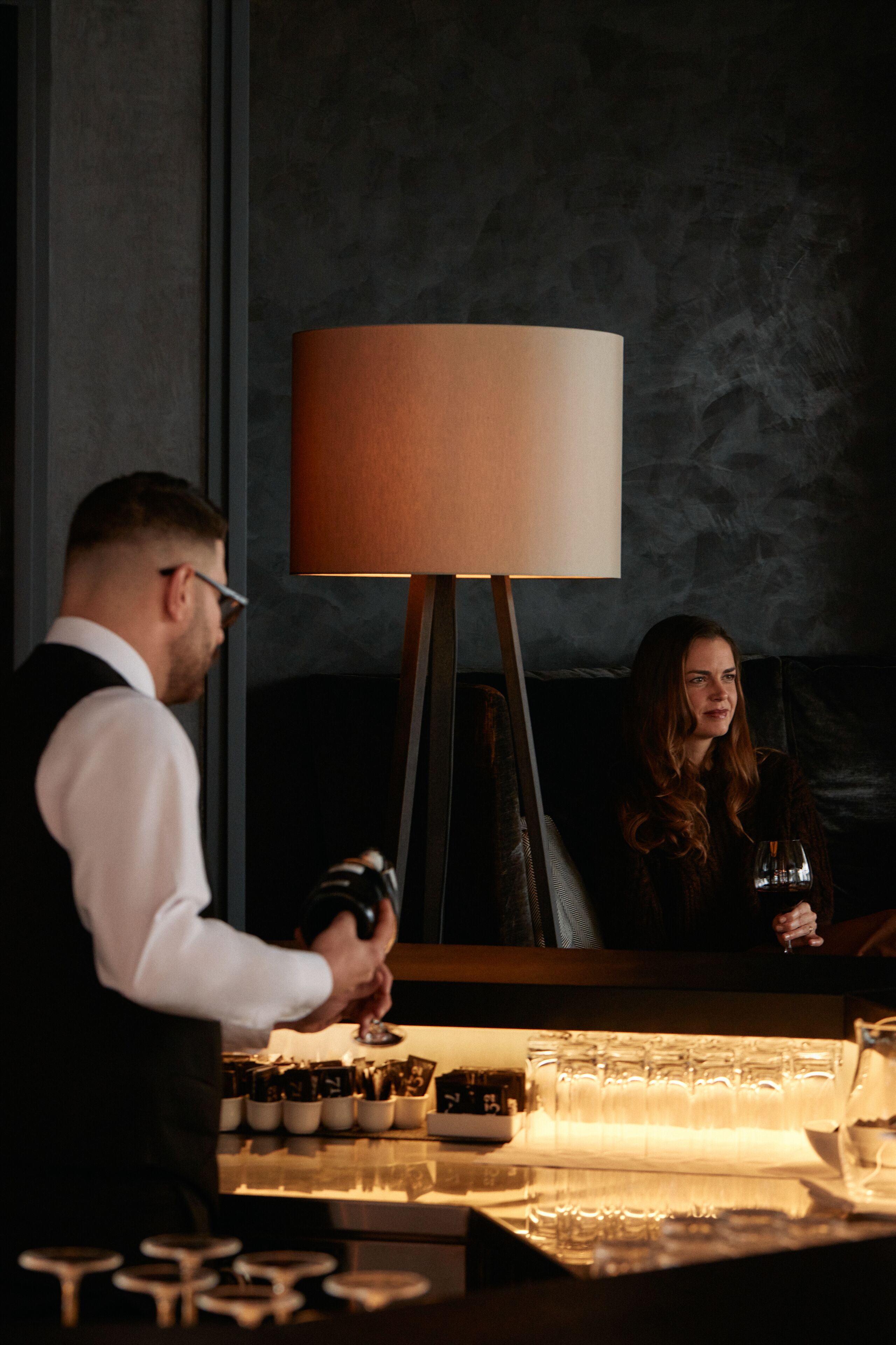A bar area with a bartender preparing drinks, a woman seated at the bar, and a large lamp in the background.