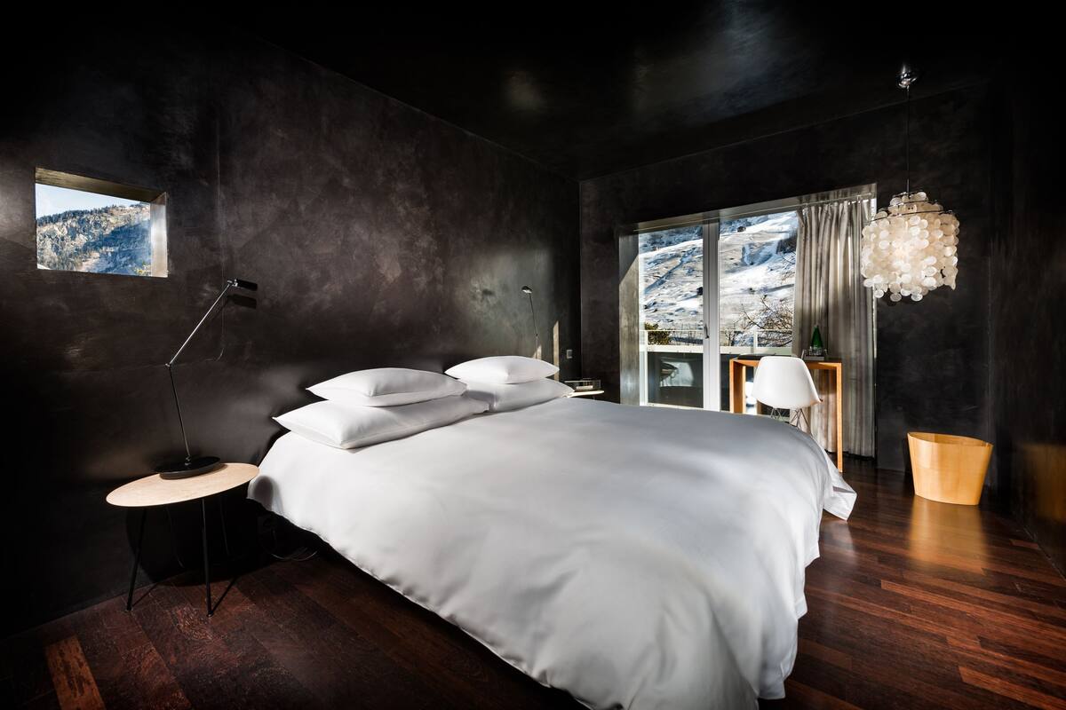 A modern bedroom with a large bed, a bedside table, a floor lamp, a window with a mountain view, and a hanging light fixture.