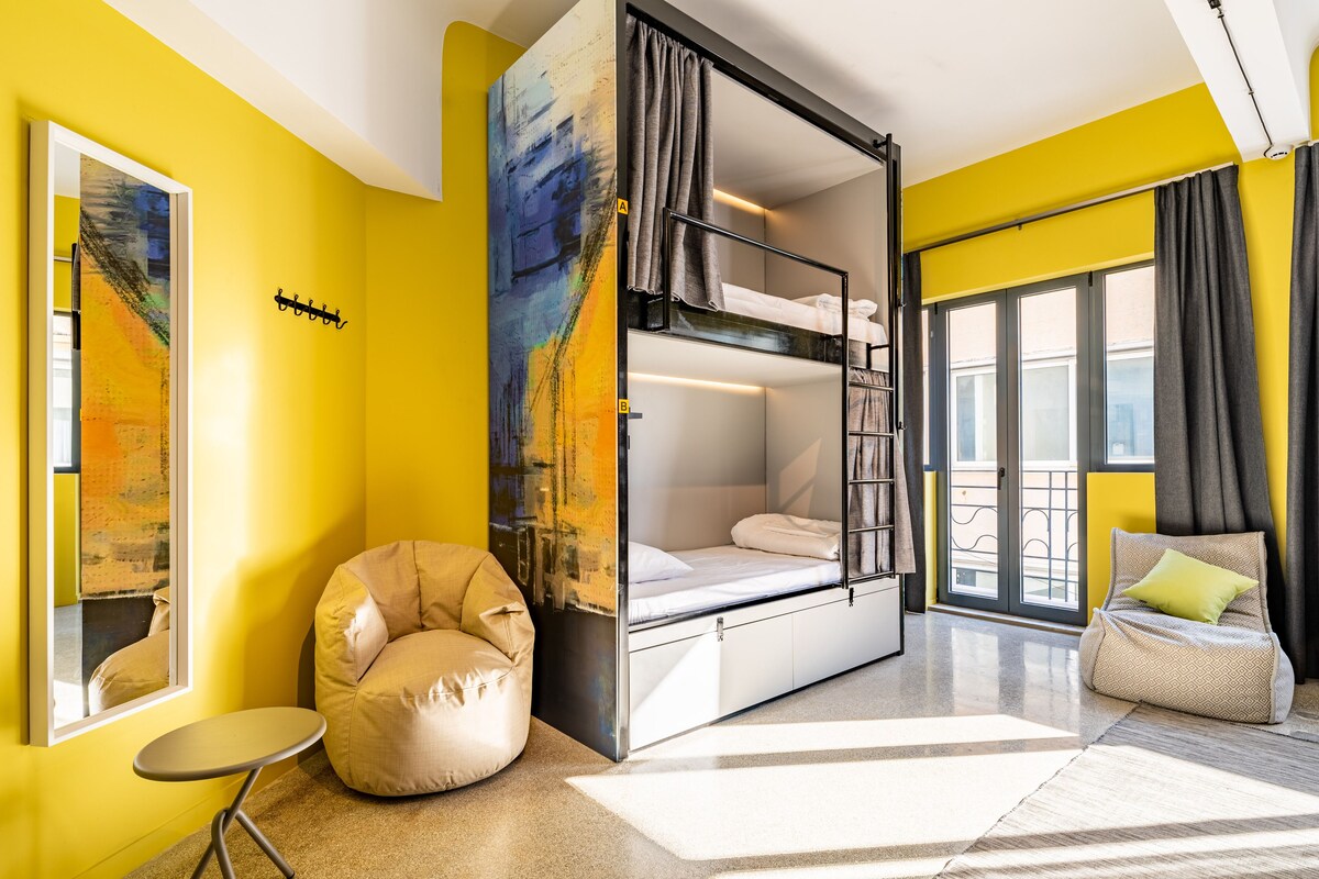 A modern hotel room with a bunk bed, a bean bag chair, a small table, and a large window with curtains.