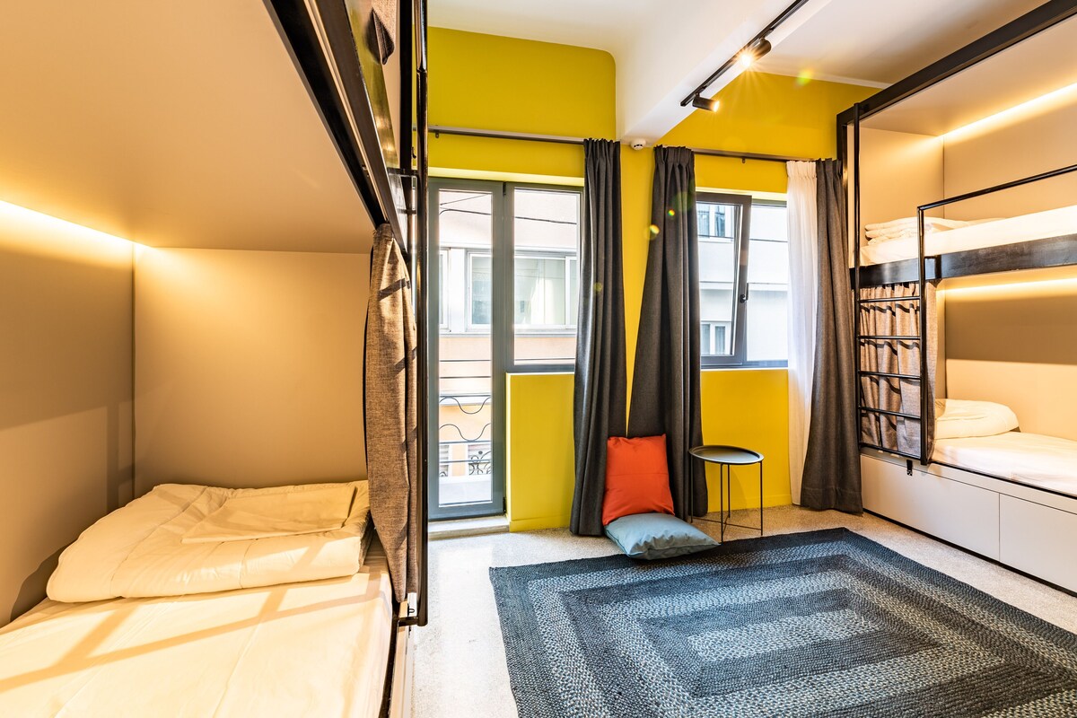A compact hotel room with a bunk bed, a small seating area, and a window with curtains.