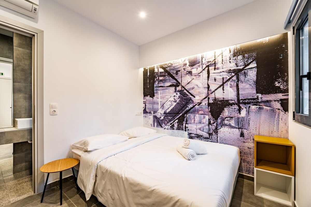 A modern hotel room with a large abstract painting on the wall, a neatly made bed, a small wooden side table, and a bathroom with a mirror and towel rack.