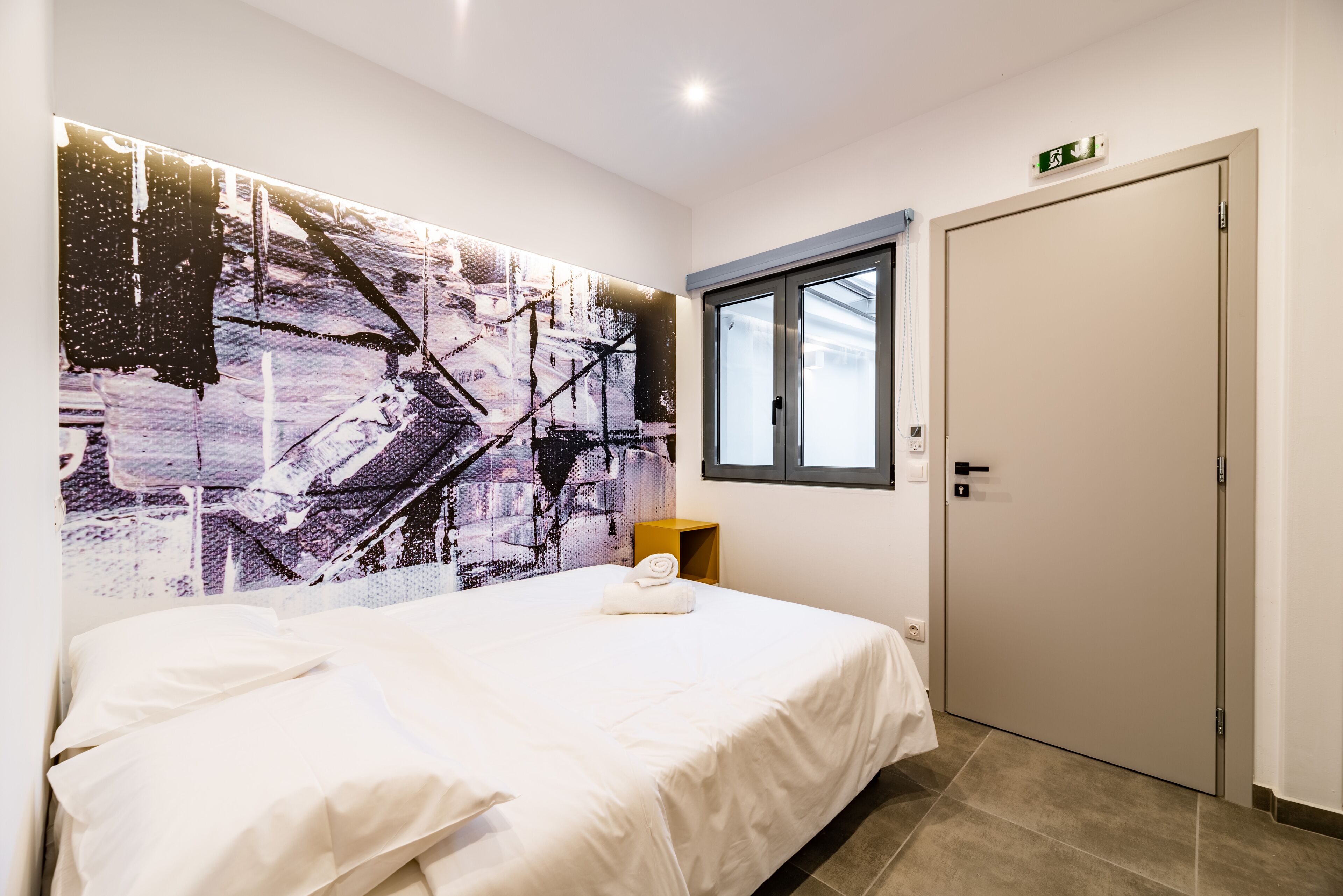 Hotellihuone, jonka seinällä on suuri abstrakti maalaus, huoneessa on valkoisilla lakanoiden peitetty sänky, ikkuna ja ovi, jossa on hätäuloskäynnin opaste.