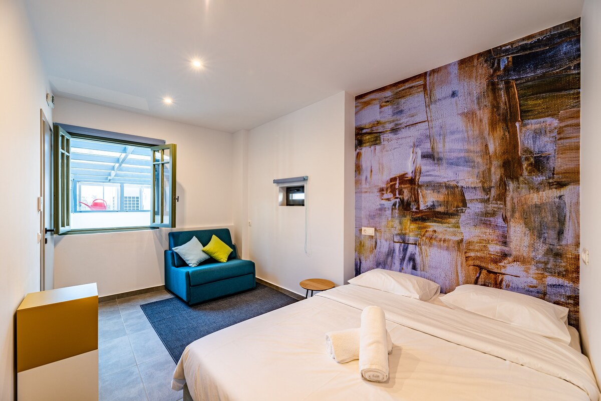 A modern hotel room with a large bed, a blue armchair, a small table, and a large abstract painting on the wall.