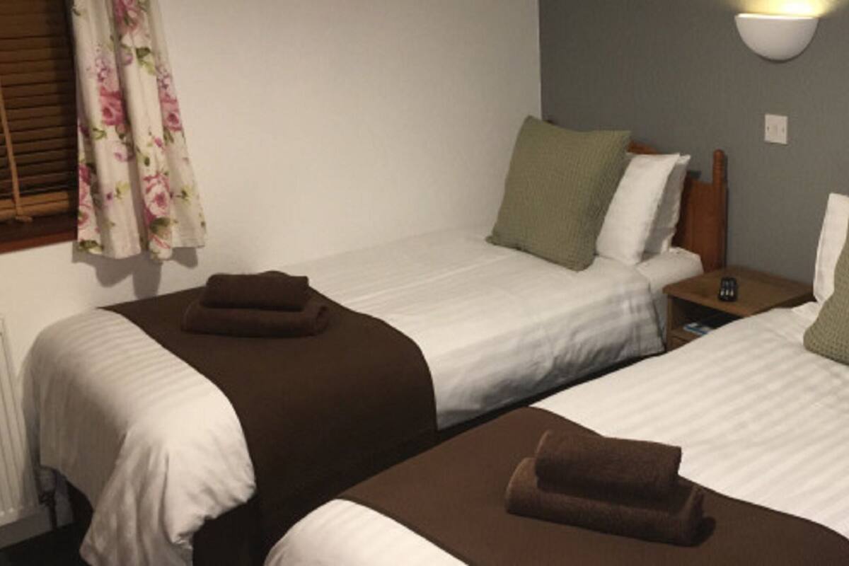 Habitación de hotel con dos camas individuales, cada una con una almohada y una manta. Hay una mesita de noche con una lámpara y un control remoto.