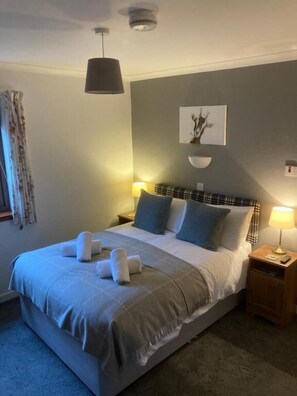 Ein ordentlich bezogenes Bett mit grauer Decke und Kissen, ein Nachttisch mit Lampe und eine Wandleuchte.