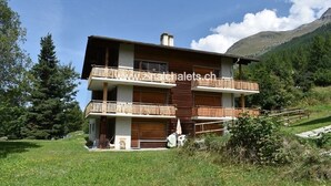 A two-story chalet with wooden balconies, set in a mountainous area with a grassy lawn and trees. - Appartment in a building in Zinal - 1 bedroom with 1 double bed (140x200) - 1 bedroom with 2 bunk be (Zinal)