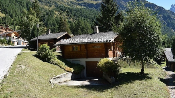 A wooden chalet with a sloped roof, surrounded by lush greenery and mountains in the background. - Individual chalet in Zinal - 1 bedroom with a double bed (140x200) - 1 bedroom with 2 beds - 1 bedro (Zinal)