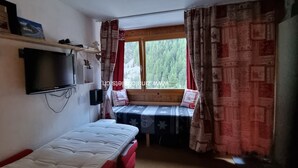 Una habitación pequeña con una cama, un televisor montado en la pared, una ventana con cortinas y un banco con mantel estampado.