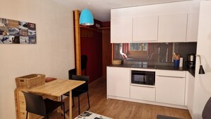 Een kleine keuken met witte kastjes, een magnetron en een koffiezetapparaat. Er staat een houten eettafel met stoelen en een mand. Boven de tafel hangt een blauwe hanglamp.