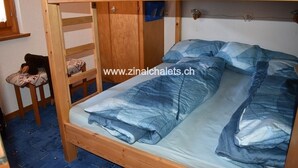 A bunk bed with a blue and white patterned bedspread and pillows, a wooden ladder, and a small chair with a patterned cushion. - Appartment in a building in Zinal - 1 bedroom with 2 beds - 1 bedroom with 2 bunk beds - living room (Zinal)