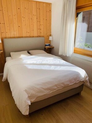 A bedroom with a wooden headboard, a bed with white bedding, a nightstand with a lamp, and a window with curtains.
