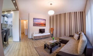 En moderne stue med sofa, sofabord og fjernsyn. Der er et køkkenområde med vask og skabe, samt et spiseområde med stole.