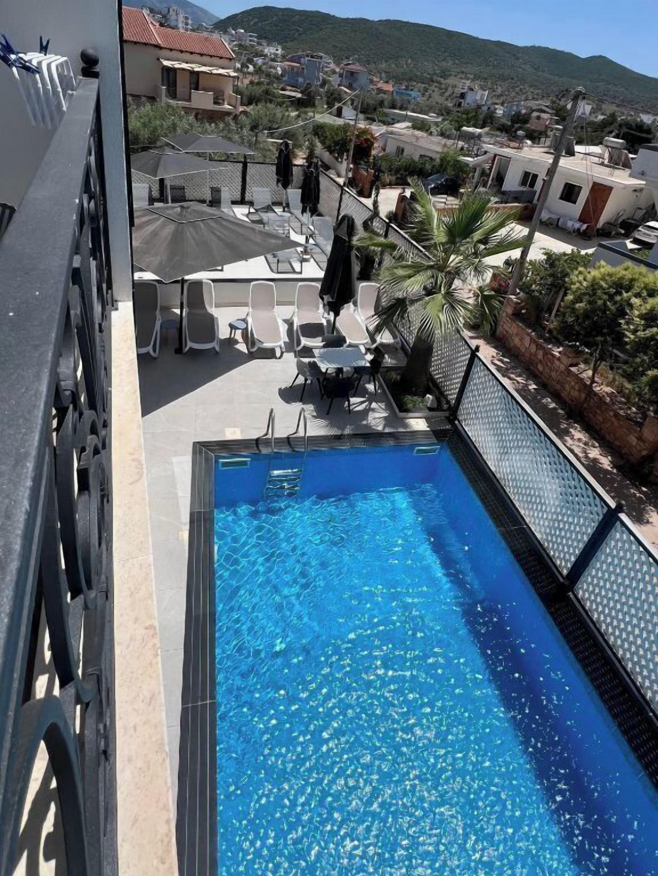 Una piscina en la azotea con agua cristalina, rodeada de sillones de baño y sombrillas, y vista a una zona residencial y colinas al fondo.