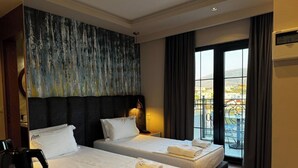 A hotel room with two beds, a large abstract painting on the wall, a view of the city through the window, and a coffee maker on the nightstand. - Area Hotel (Ksamil)