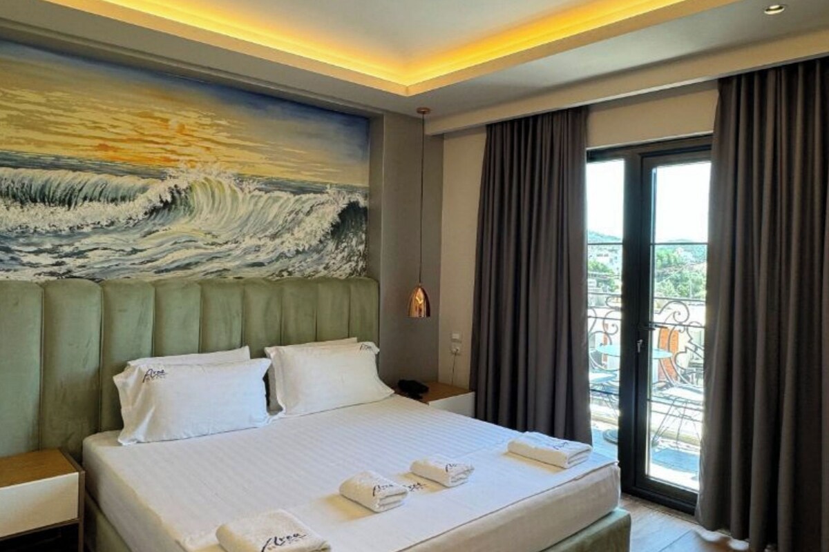 A hotel room with a large bed, a painting of waves on the wall, a balcony with curtains, and a wooden floor.