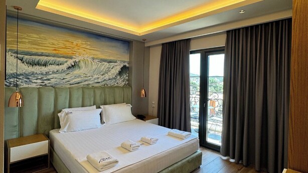 A hotel room with a large bed, a painting of waves on the wall, a balcony with curtains, and a wooden floor. - Area Hotel (Ksamil)