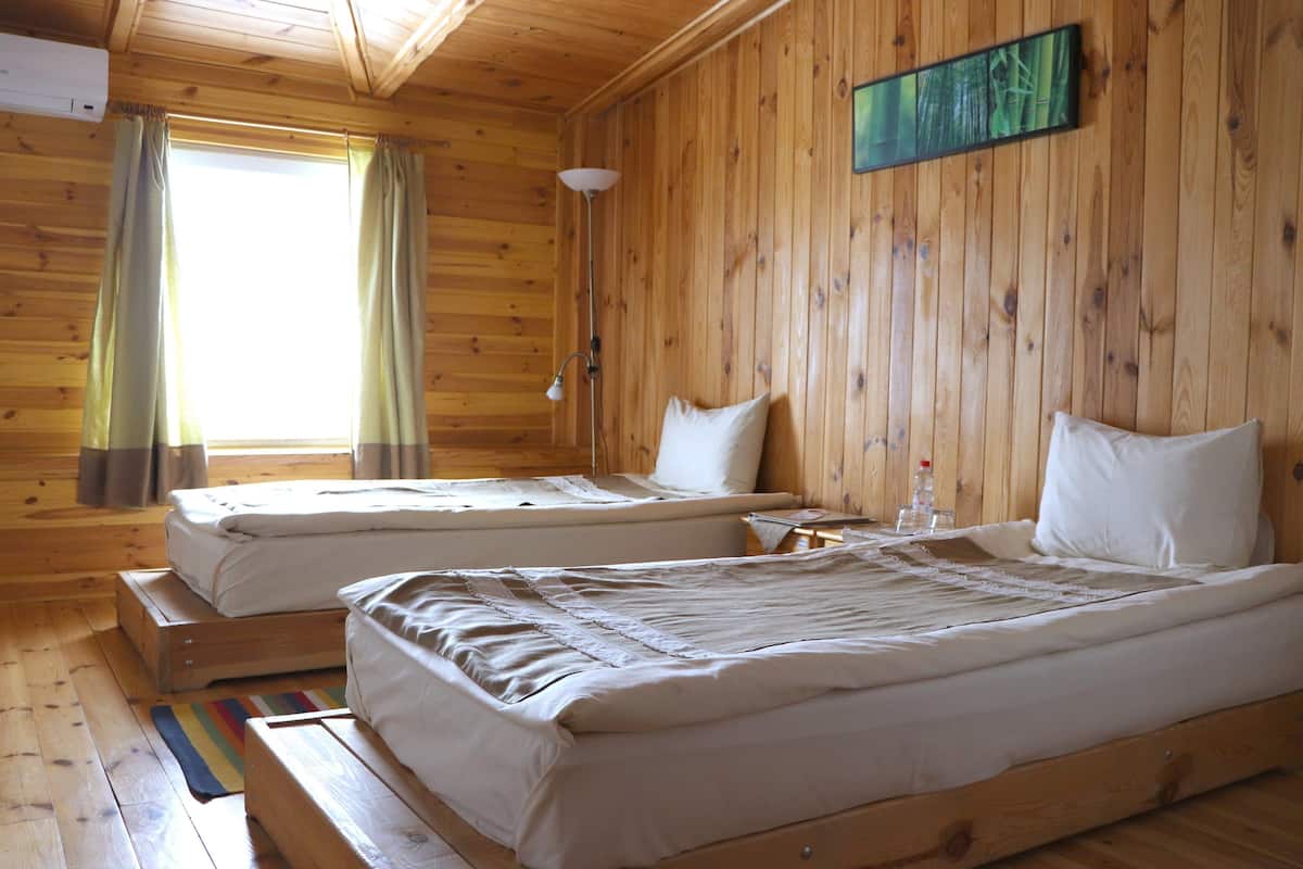 A wooden cabin room with two beds, a window with curtains, and a painting on the wall.