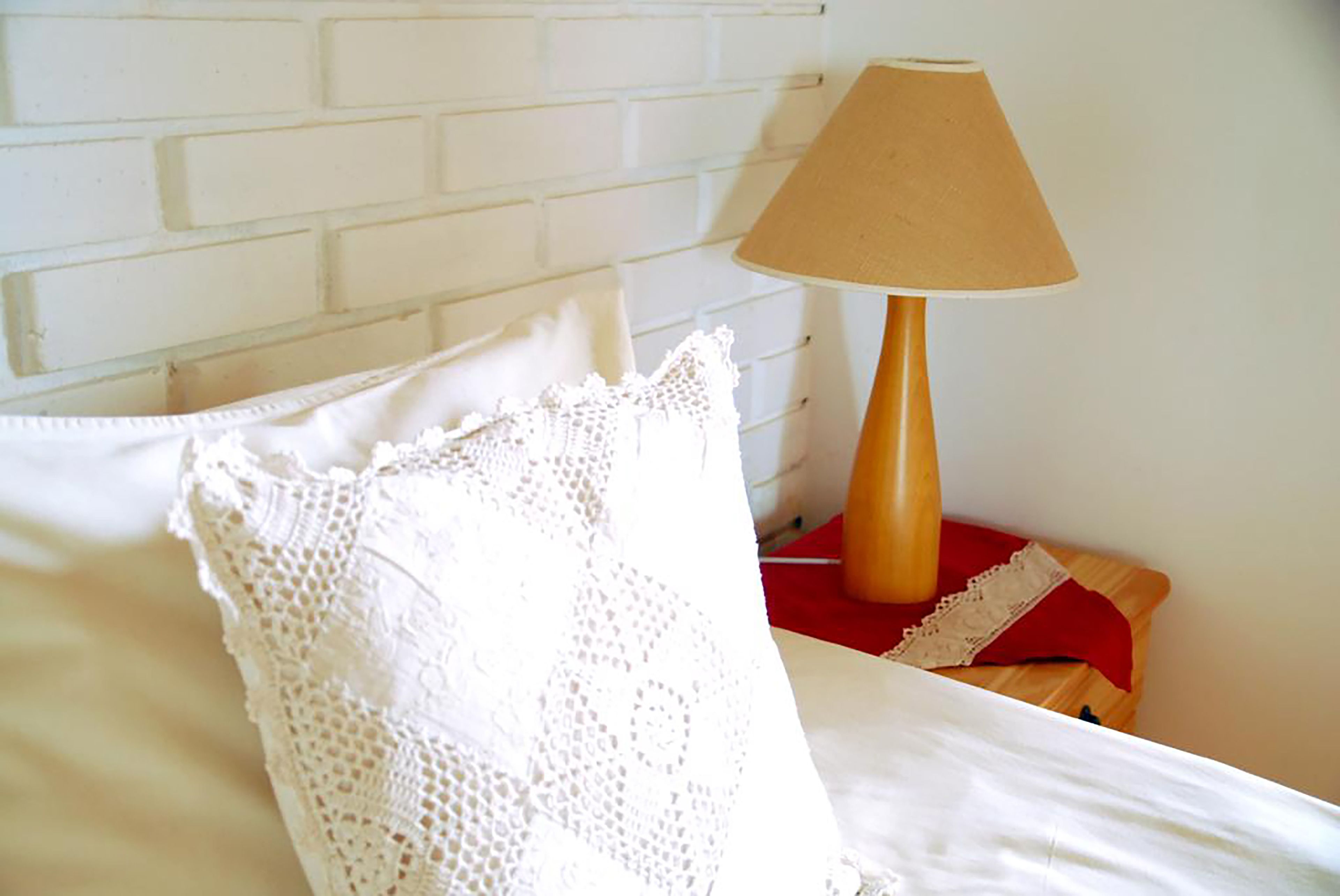 A neatly made bed with a white pillow and a white lace throw, a wooden bedside table with a lamp, and a white brick wall.