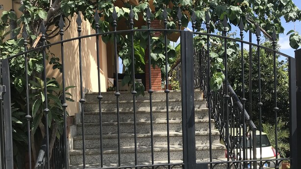 Um portão de metal com um muro de pedra ao fundo, céu claro e alguma vegetação.