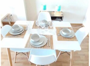 Mesa de comedor dispuesta con platos, tazones y cubiertos blancos, rodeada de sillas blancas.
