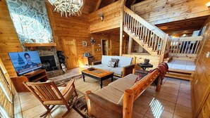 A cozy log cabin interior with a stone fireplace, wooden furniture, and a television displaying a news channel. - Spacious 5BR Mountain Lodge | Hot Tub, Pool Table & Scenic Views (Grassy Creek)