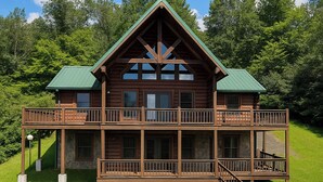 A two-story log cabin with a balcony on the upper level, surrounded by lush greenery. - Spacious 5BR Mountain Lodge | Hot Tub, Pool Table & Scenic Views (Grassy Creek)