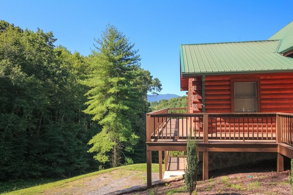 A log cabin with a wooden deck and a green roof, surrounded by dense forest and mountains in the background. - Spacious 5BR Mountain Lodge | Hot Tub, Pool Table & Scenic Views (Grassy Creek)