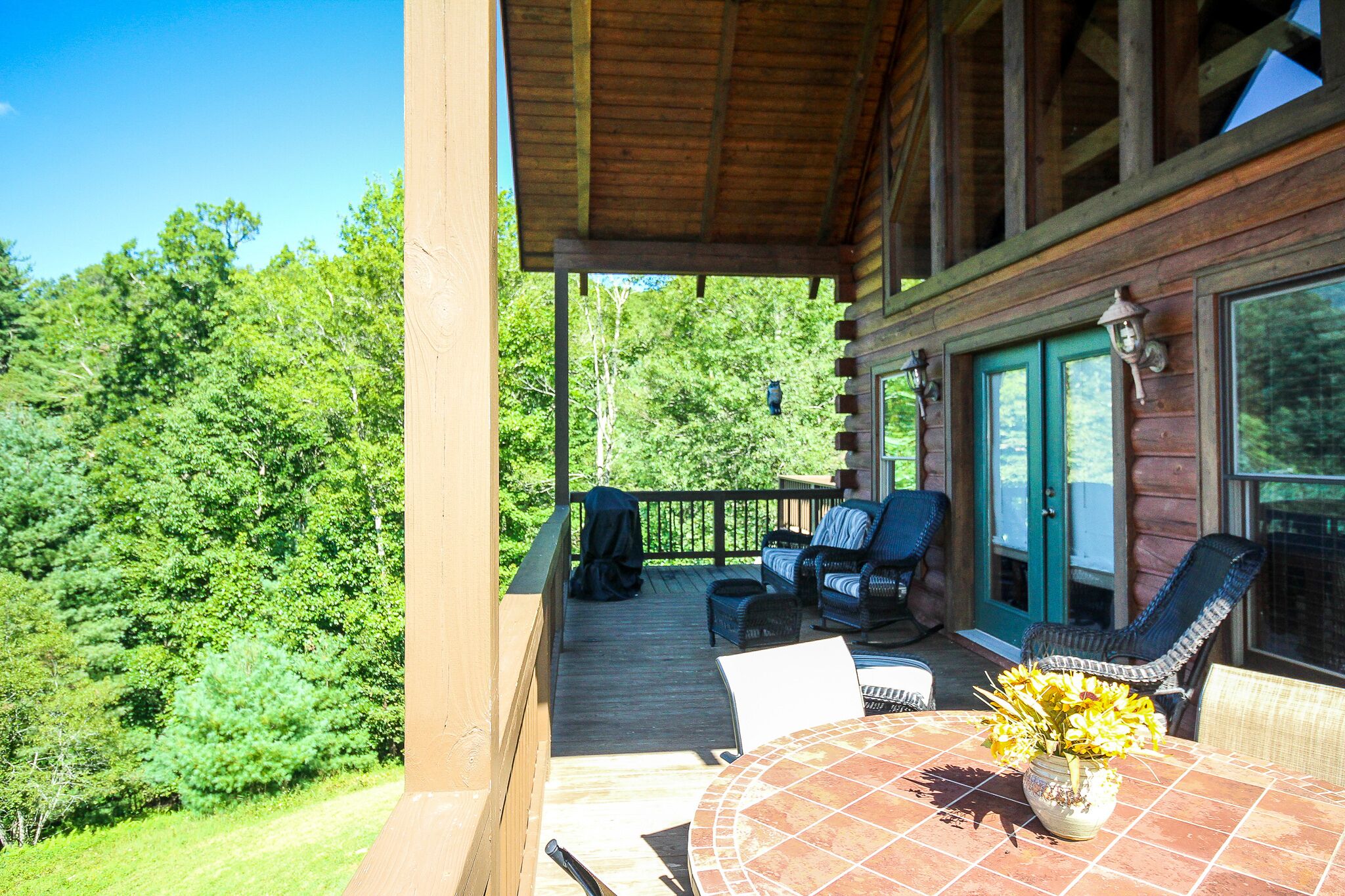 A wooden deck with outdoor furniture, a table with a vase of flowers, and a view of a forested area.