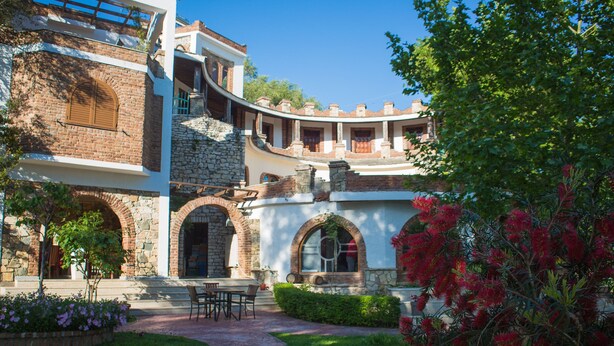 A courtyard with a stone wall, a wooden bench, and a pathway leading to a building with arched windows and a balcony. - Hotel Livia (Xarrë)