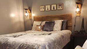 Un dormitorio con cabecera de madera, cama con colcha estampada, dos lámparas de pared y una pequeña decoración de la Torre Eiffel sobre la mesita de noche.
