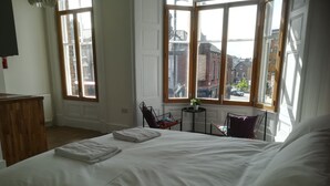 A bedroom with a large bed, two chairs, a small table with a plant, and large windows with a city view. - McMillan House Studio Apartments (York)