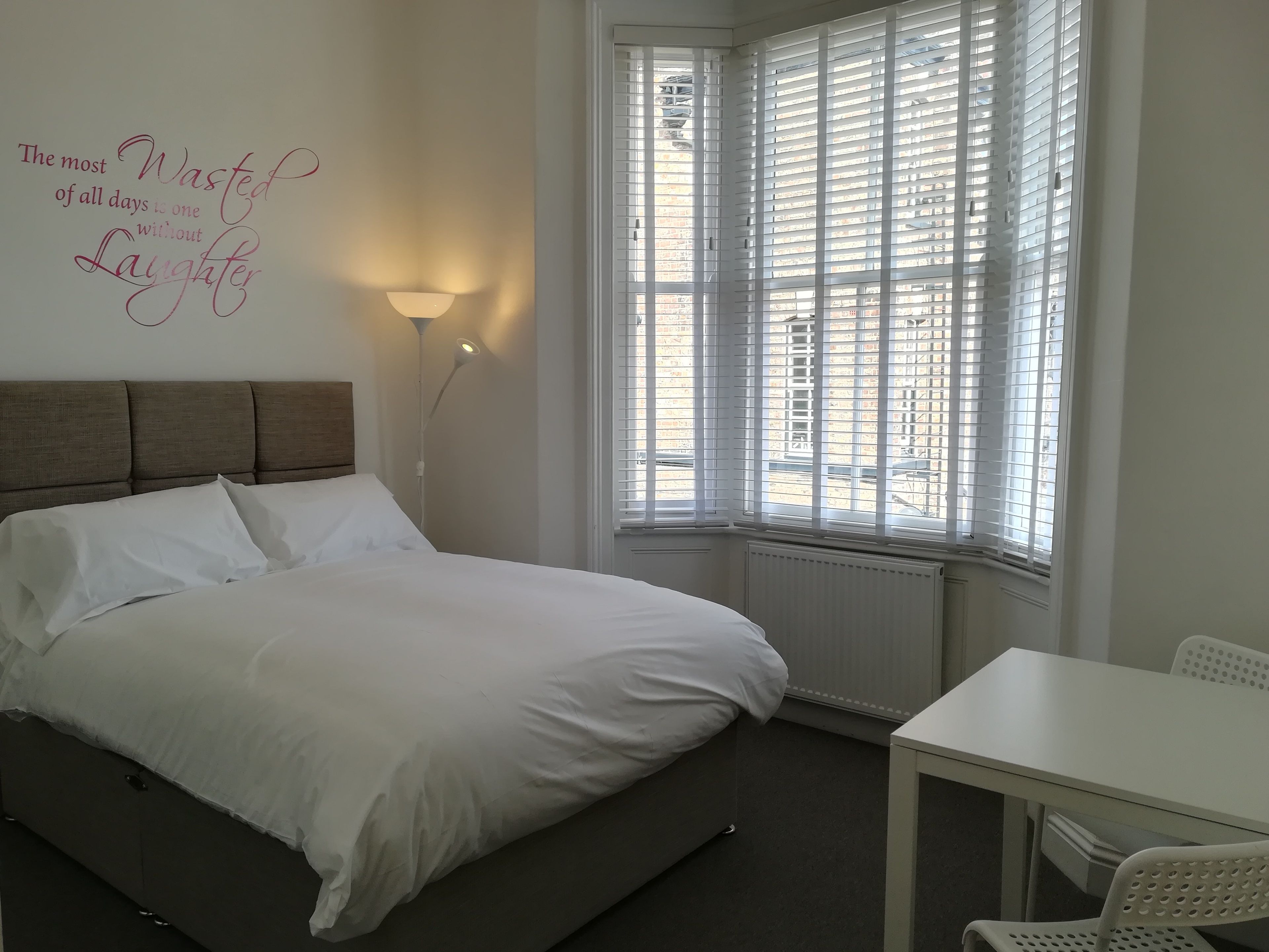 A neatly made bed in a room with a large window, a wall-mounted lamp, and a small table.