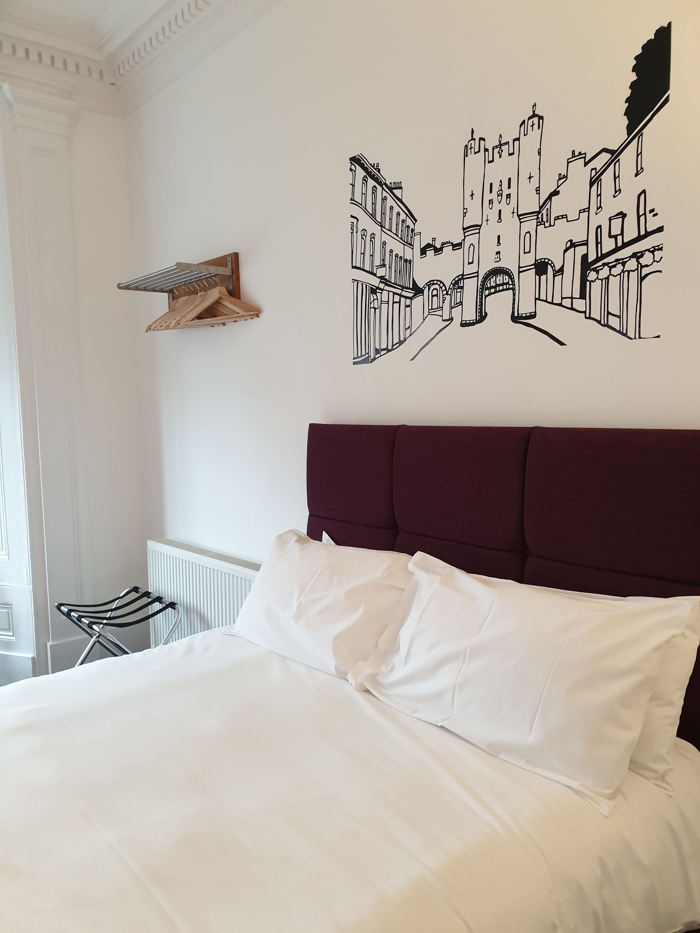 A bedroom with a large bed, a wooden drying rack, and a wall mural of a cityscape.