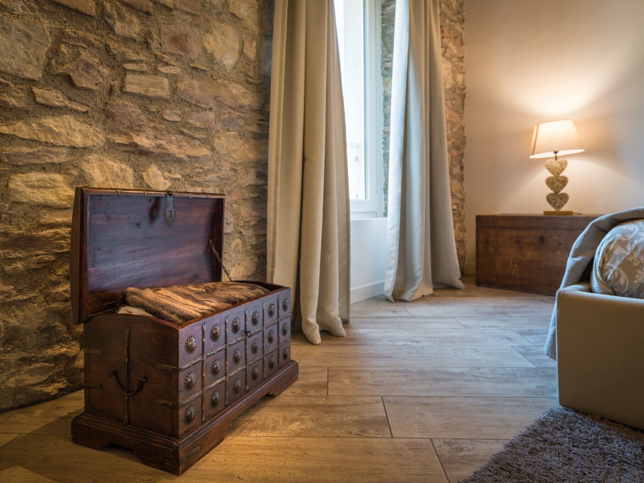 A room with a stone wall, a wooden chest, a window with curtains, a lamp, and a sofa.