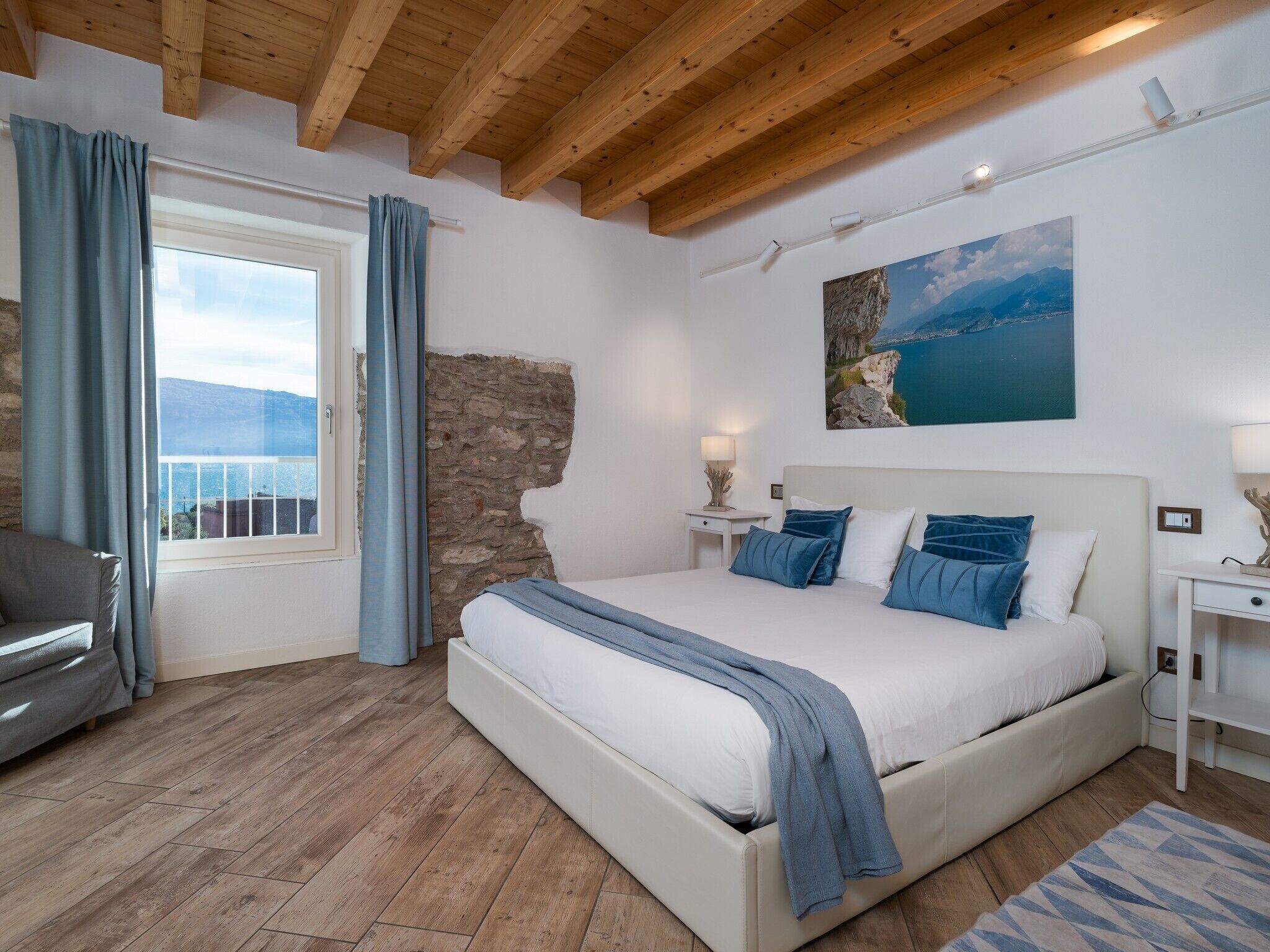 A bedroom with a large bed, a view of the sea through a window, and a stone wall.