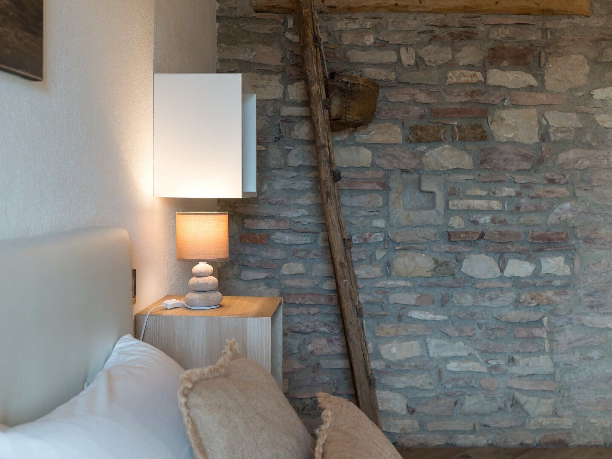 A bedroom with a stone wall, a bed with white linens, a bedside table with a lamp, and a wooden beam.