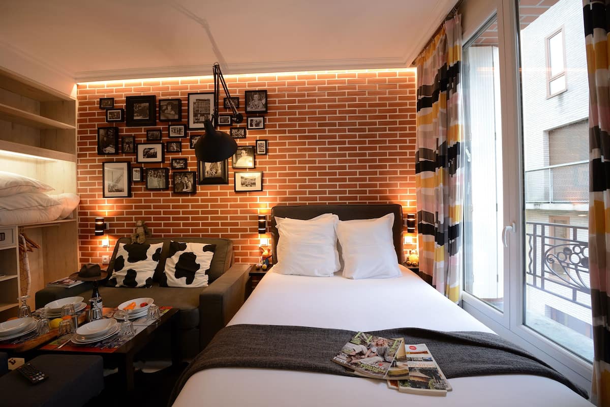 A hotel room with a brick wall, a bed with white bedding, a nightstand with a lamp, a sofa with a cow print pillow, and a window with curtains.