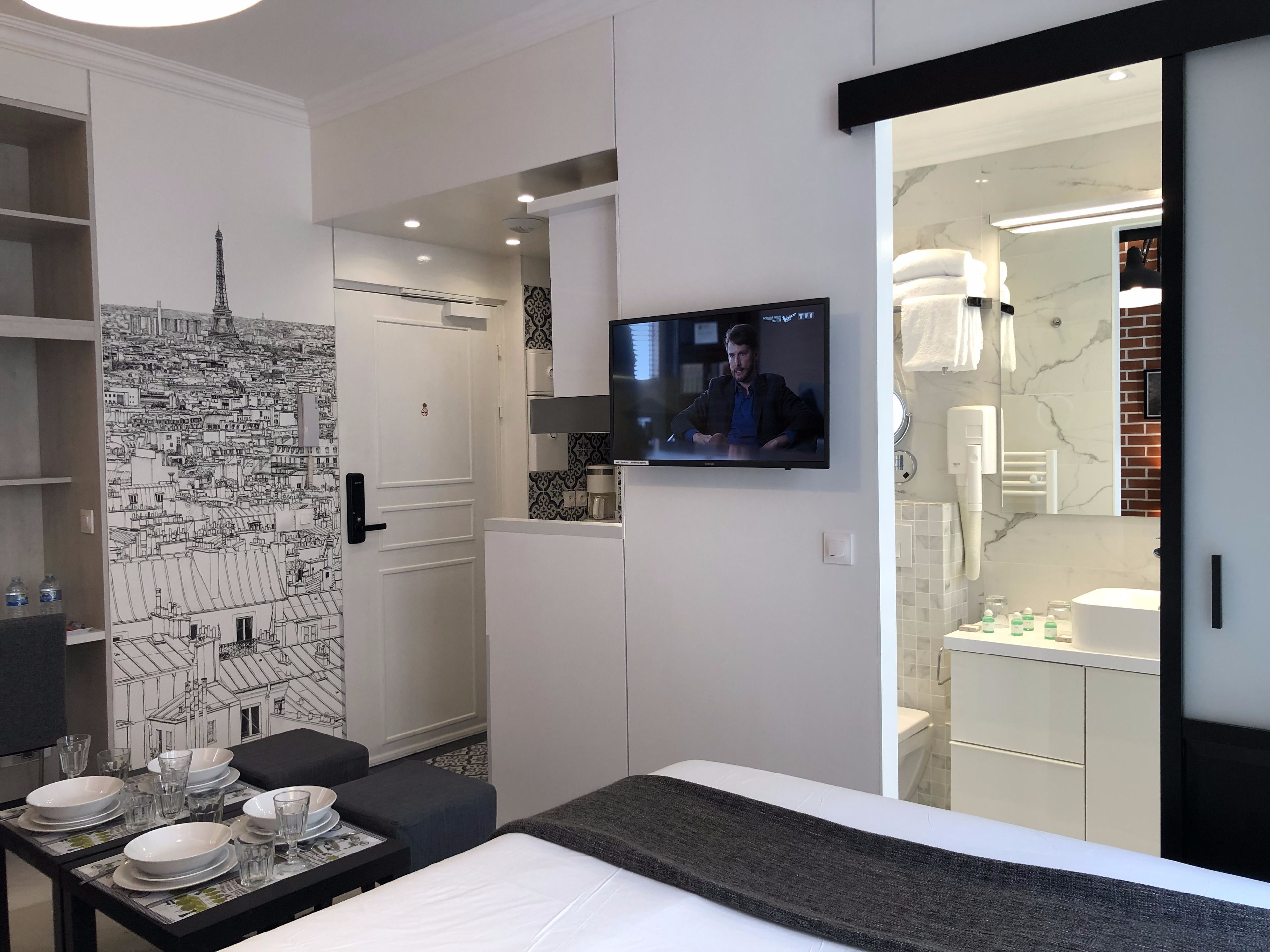 Une chambre d’hôtel moderne avec une fresque représentant un paysage urbain, un téléviseur à écran plat, un coin repas avec une table dressée pour deux personnes, et une salle de bain avec un mur en marbre.
