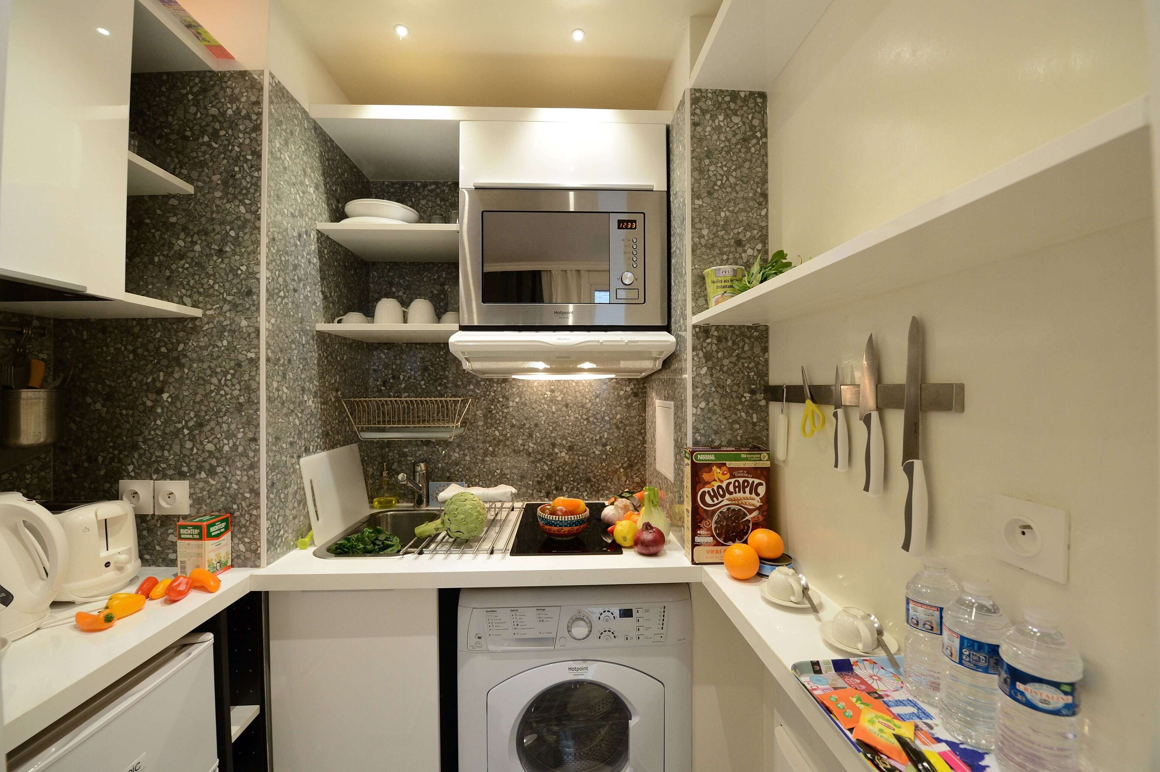Cocina compacta con microondas, lavavajillas y fregadero. Hay verduras, una caja de Chocopice y botellas de agua en la encimera.