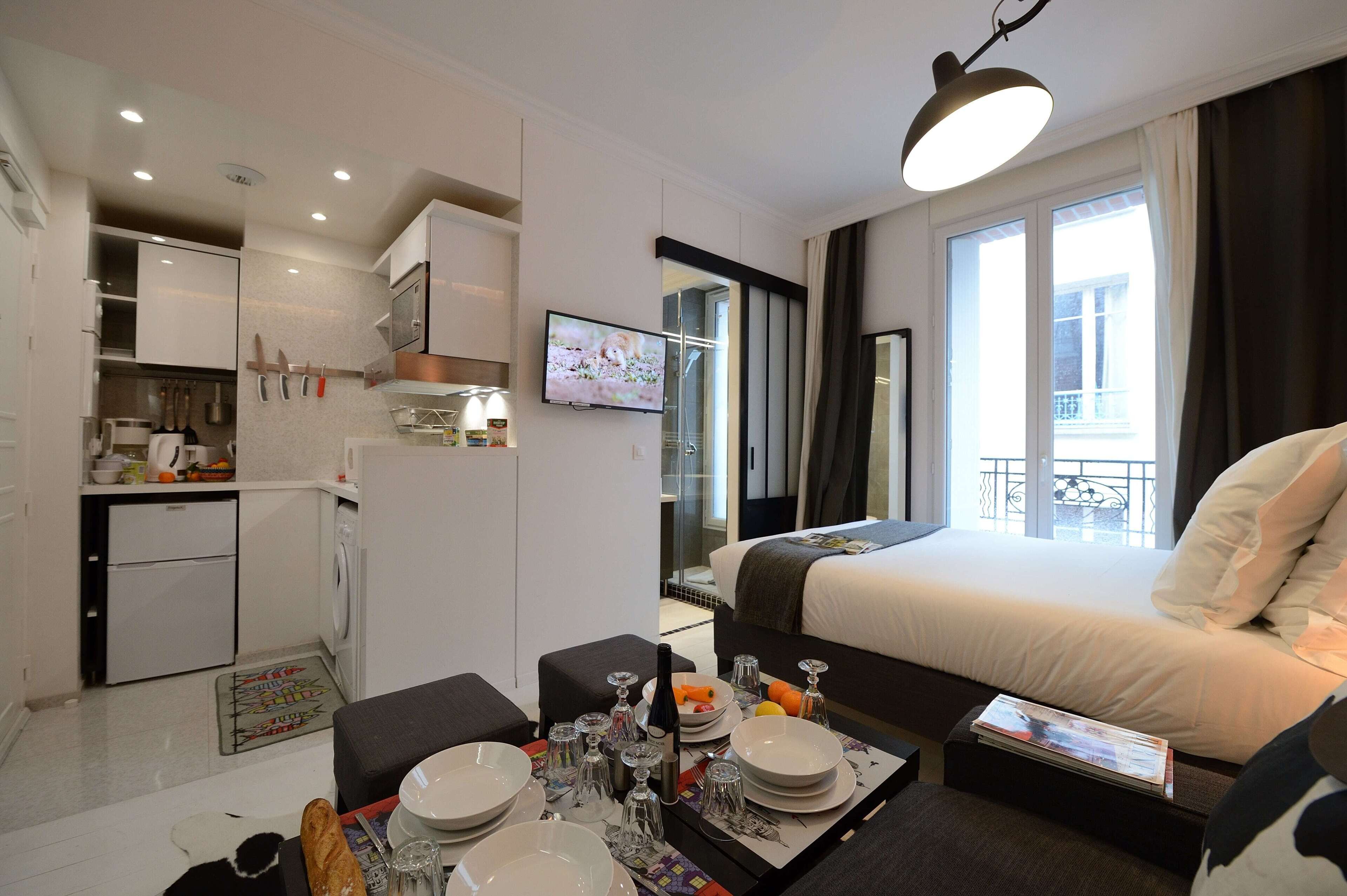 A hotel room with a bed, a dining area with a table set for two, a kitchenette, and a large window with a view of the outside.