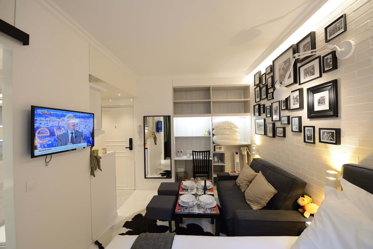A modern hotel room with a flat-screen TV, a sofa, a dining table set, and a wall with framed photos.