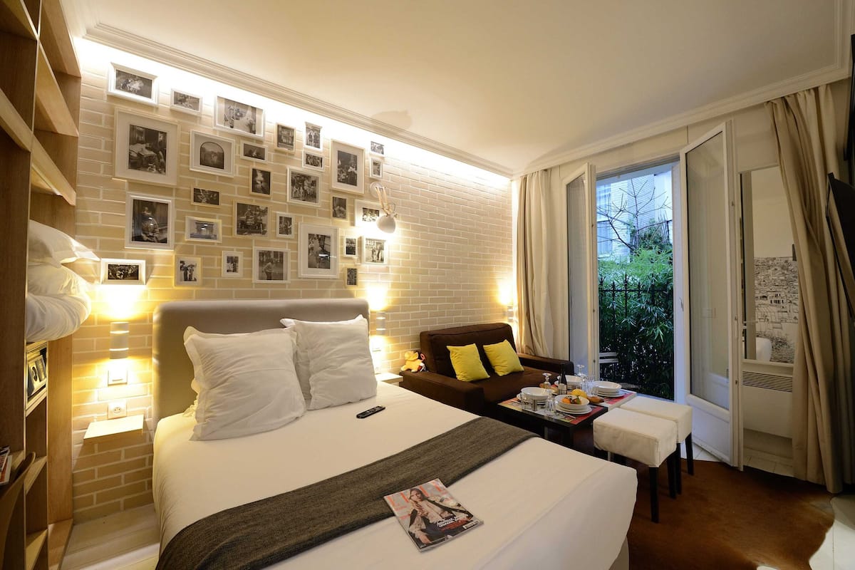 A hotel room with a neatly made bed, a bedside table with a lamp, a small sofa with cushions, a window with curtains, and a wall decorated with framed photos.