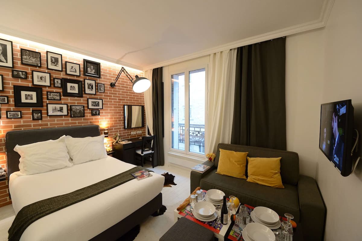 A hotel room with a bed, a sofa, a dining table set with plates and glasses, a TV, and a wall with framed pictures.