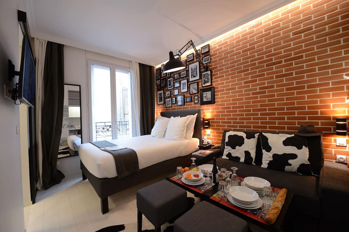 A modern hotel room with a large bed, a dining area with a table set for two, and a brick wall with framed pictures.