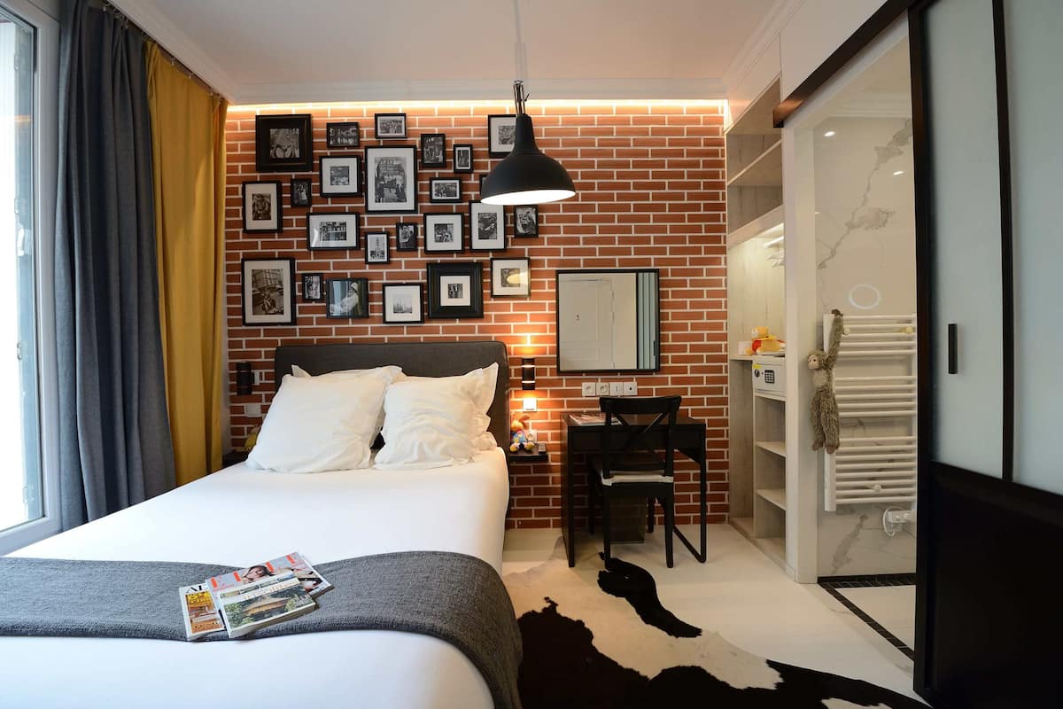 A hotel room with a bed, a desk, a chair, a black lamp, a wall with framed pictures, and a white door.