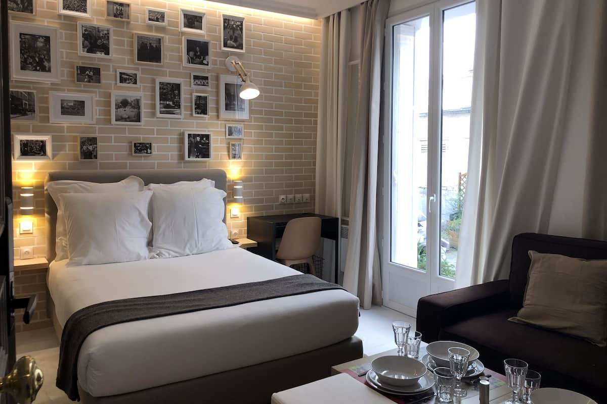 A hotel room with a large bed, a desk with a chair, a sofa, and a dining table set for two. There are framed pictures on the wall and a window with curtains.