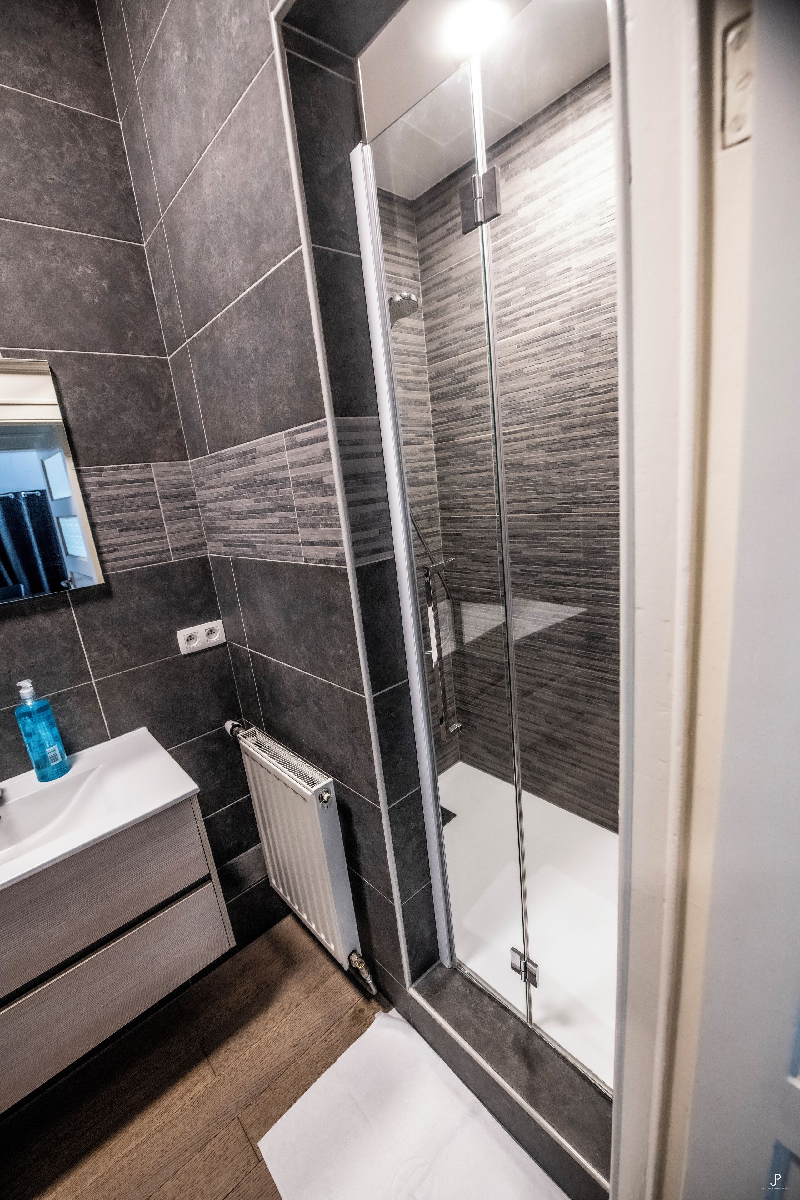 A modern bathroom with a glass-enclosed shower, dark tiles, a white sink, and a mirror.