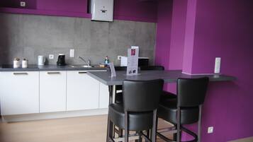Una cocina moderna con armarios blancos, encimera gris y pared morada. Hay dos sillas negras junto a una pequeña mesa.