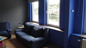 A room with a blue sofa, a small table, and a window with a view of a building and water. - L'Inattendu sur la Croisette Dinant Centre (Dinant)